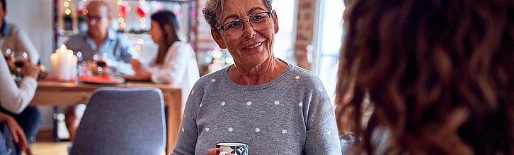 Frau mit Kaffeetasse in der Hand im Gespräch, im Hintergrund Menschen an einem Tisch